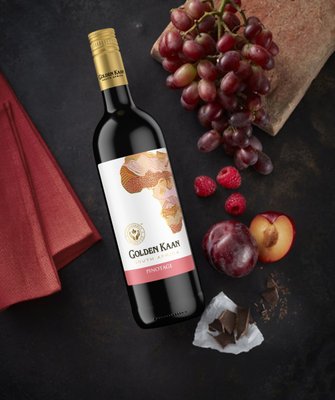 Eine Flasche Golden Kaan Pinotage Wein steht auf einem dunklen Tisch, umgeben von Trauben, Himbeeren und einer Pflaume.