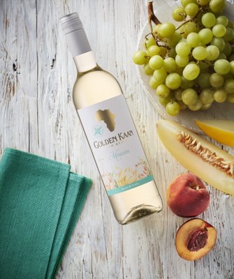 Auf einem Holzuntergrund liegt eine Flasche Golden Kaan Moscato, umgeben von saftigen Trauben und frischem Obst.