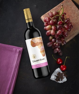 Auf einem dunklen Tisch liegt eine Flasche Golden Kaan Merlot, umgeben von Trauben, Kirschen und einem Stück Schokolade.