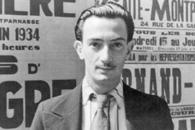 Salvador Dali mit einem schmalen Gesicht und einem Schnurrbart steht vor einer Wand voller Plakate aus den 1930er Jahren.