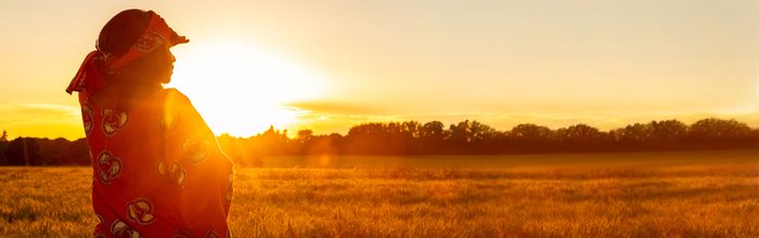 Eine Person in traditioneller Kleidung steht in einem goldenen Feld und blickt auf den Sonnenuntergang am Horizont.